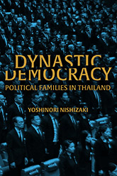 Gold letters spell out 'Dynastic Democracy.' 'Dynastic' eclipses a small portion of the top side of 'Democracy' while 'Democracy' blots out a small portion of the bottom part of 'Dynastic.' In an auditorium, a multitude of politicians stand attention in the background.